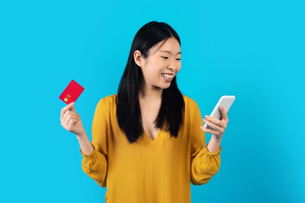 Alegre mujer asiática con teléfono inteligente y tarjeta bancaria en azul