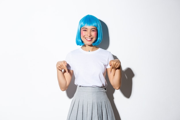 Alegre mujer asiática en traje de estilo anime, con peluca azul y sonriendo feliz, señalando con el dedo hacia abajo, mostrando publicidad, de pie.