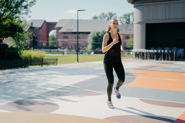 Alegre mujer atlética corre activamente en el estadio vestida con ropa deportiva negra y disfruta de actividades físicas al aire libre durante el día de verano lleno de energía. Concepto de deporte y motivación de personas.