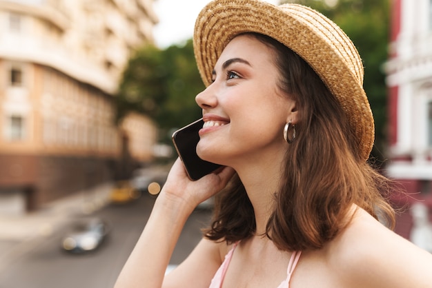 Alegre mujer bonita joven feliz sonriente positiva con hermoso sombrero caminando por la calle hablando por teléfono móvil.