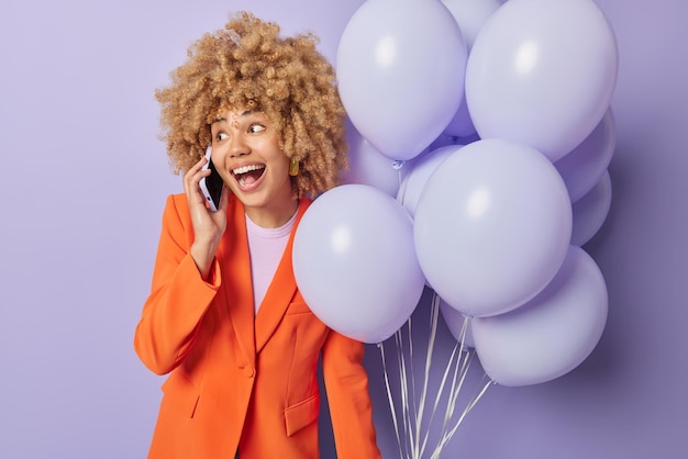 Alegre mujer de cabello rizado vestida con ropa de moda hace una llamada telefónica sostiene un montón de globos inflados invita a amigos a una fiesta aislada sobre fondo púrpura Concepto de ocasión festiva