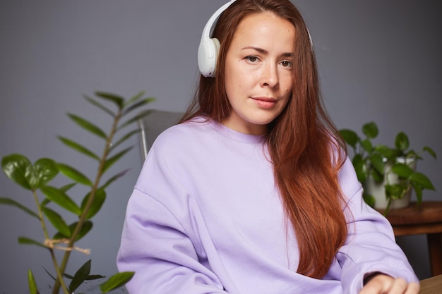 Alegre mujer caucásica escuchando música o podcast Muchas plantas verdes alrededor Mujer pelirroja con suéter violeta