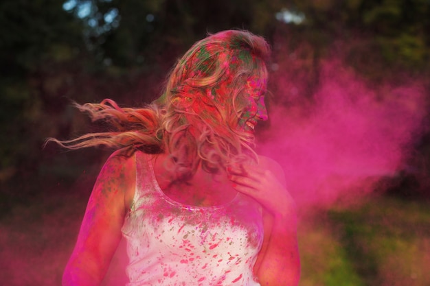 Alegre mujer caucásica iwith pelo rizado de pie en una nube de pintura seca rosa, celebrando el festival de Holi