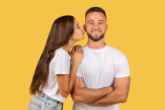 Alegre mujer europea del milenio con camiseta blanca besa a un hombre fuerte en la mejilla disfruta de una cita romántica