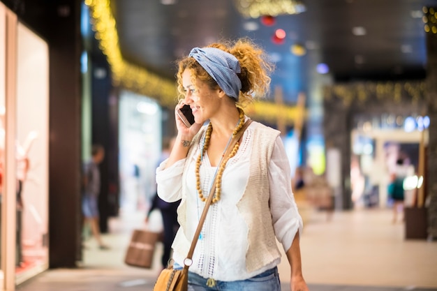 Alegre mujer feliz hacer compras en el centro comercial mientras llama con el teléfono y mira las tiendas para decidir qué comprar y usar para estar a la moda y hermosa - dama moderna urbana
