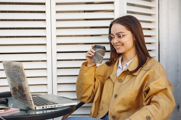 Alegre mujer independiente bebiendo café en un café de la calle mientras trabaja en una computadora portátil en un proyecto remoto