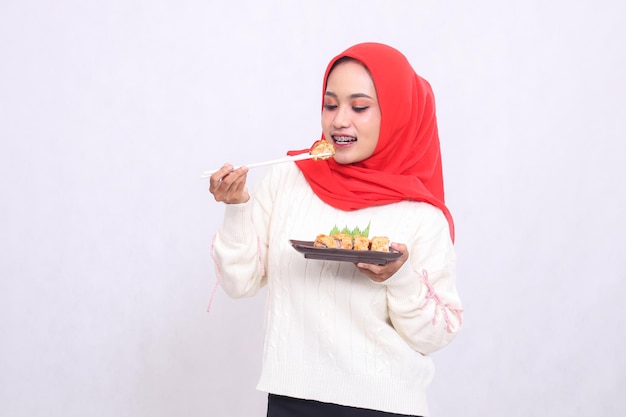 Una alegre mujer indonesia con hijab sostiene palillos para comer en el plato y lleva un plato que contiene sushi