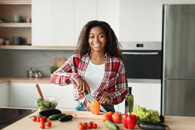 Alegre mujer joven bastante negra en cortes casuales verduras orgánicas para ensalada en el interior de la cocina moderna