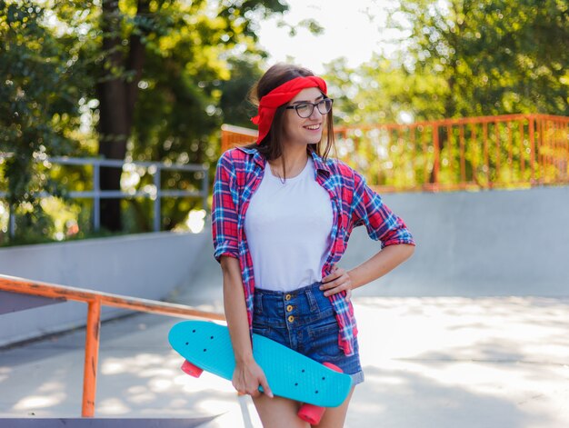 Alegre mujer joven inconformista emocional vestida con ropa elegante tiene patineta en su mano en skatepark en día soleado