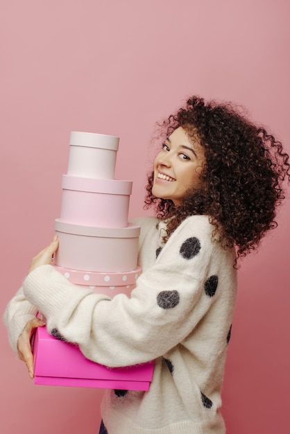 Alegre mujer joven de piel clara abrazando una pila de cajas de regalo de su familia en el interior La dama de cabello oscuro y rizado sonríe con los dientes vestida con un suéter blanco Concepto de estado de ánimo festivo