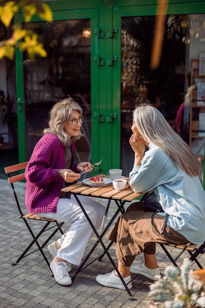 Alegre mujer madura con un amigo mesa pequeña con postre y bebidas al aire libre