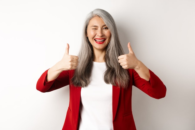 Alegre mujer madura asiática guiñando un ojo, sonriendo complacida y mostrando el pulgar hacia arriba en señal de aprobación, como y de acuerdo, de pie contra el fondo blanco.