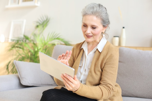 Alegre mujer de mediana edad sentada en el sofá, usando aplicaciones de tableta de computadora, mirando la pantalla, leyendo buenas noticias en la red social, comprando o charlando en línea.