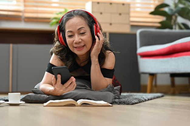 Alegre mujer de mediana edad usando auriculares usando un teléfono móvil mientras está acostada en el suelo en casa