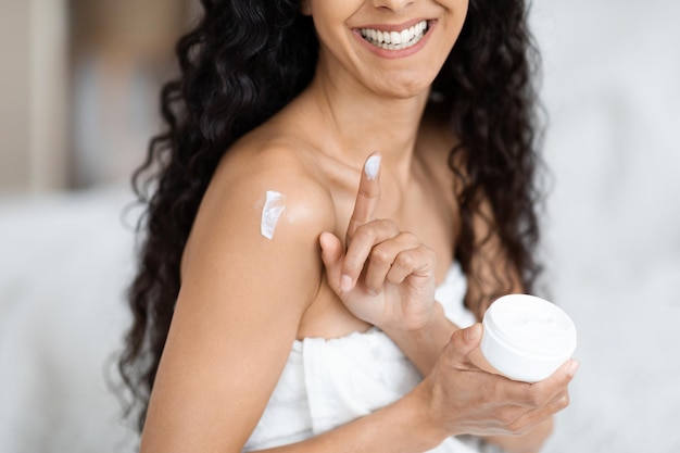 Foto alegre mujer milenaria árabe con el pelo largo y rizado en ropa doméstica aplicando crema en el hombro