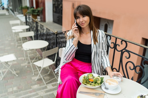 Alegre mujer morena bebiendo café, usando el teléfono inteligente y disfrutando del desayuno en un elegante café en la calle.