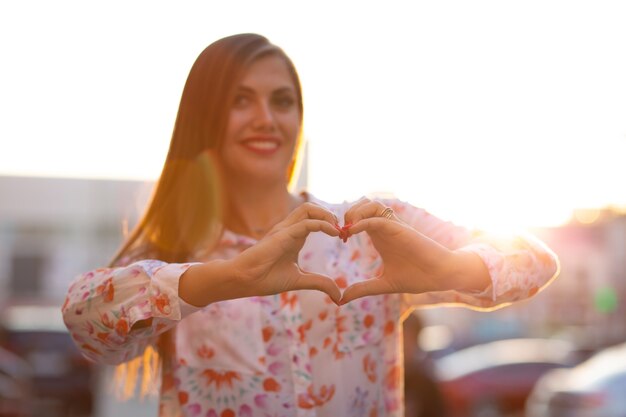 Foto alegre mujer morena con cabello largo sosteniendo las manos en forma de corazón en la calle con rayos de sol