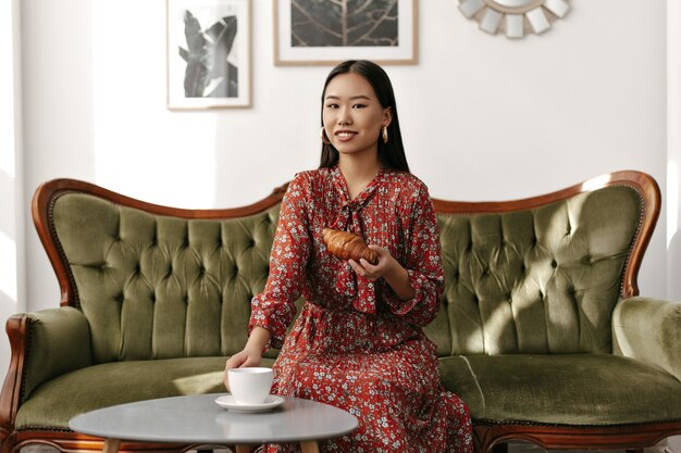 Alegre mujer morena en vestido floral rojo se sienta en un sofá suave de terciopelo verde, sostiene un sabroso croissant y una taza de té