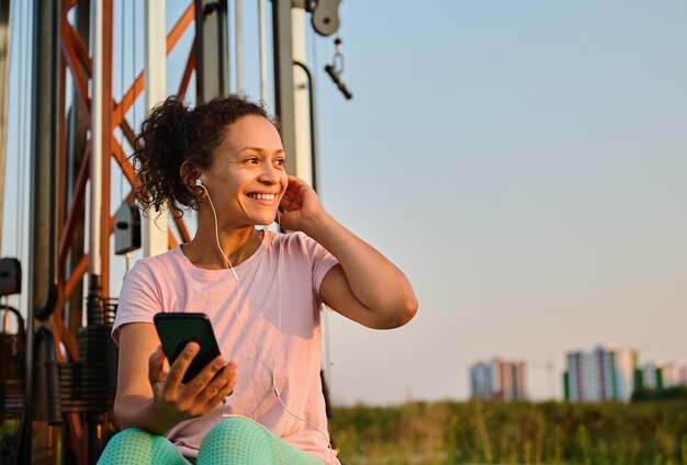 Alegre mujer multiétnica en ropa deportiva con auriculares escuchando música sonríe mirando a un lado mientras descansa entre conjuntos durante el entrenamiento al aire libre en el campo deportivo