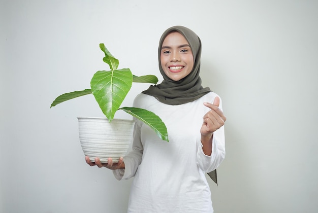 Una alegre mujer musulmana asiática con camiseta y hiyab sosteniendo una planta aislada de fondo blanco