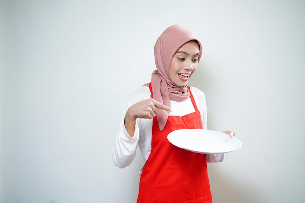 Alegre mujer musulmana asiática en delantal rojo apuntando al plato vacío Anuncio de comida Concepto de cocina