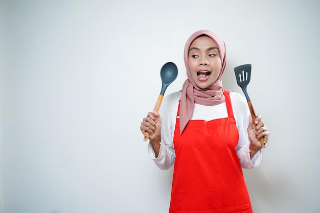 Alegre mujer musulmana asiática en delantal rojo con cuchara de sopa y espátula Utensilios de cocina Concepto de cocina