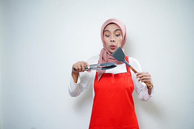 Alegre mujer musulmana asiática en delantal rojo con espátula y pinzas Utensilios de cocina Concepto de cocina