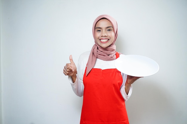 Alegre mujer musulmana asiática en delantal rojo con plato vacío Anuncio de comida Concepto de cocina