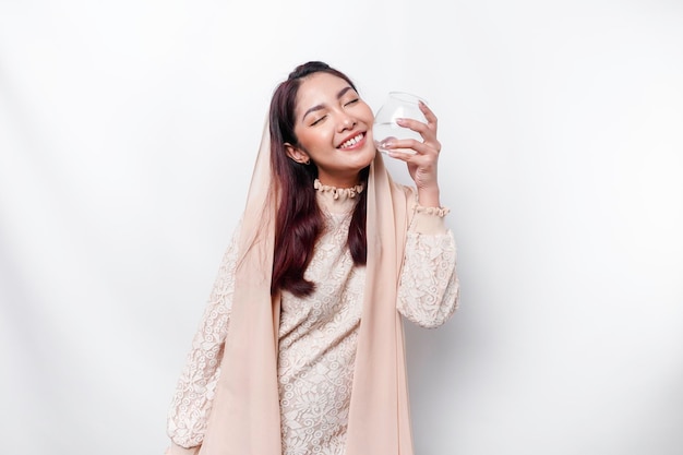 Una alegre mujer musulmana asiática con pañuelo en la cabeza está bebiendo un vaso de agua aislado de fondo blanco