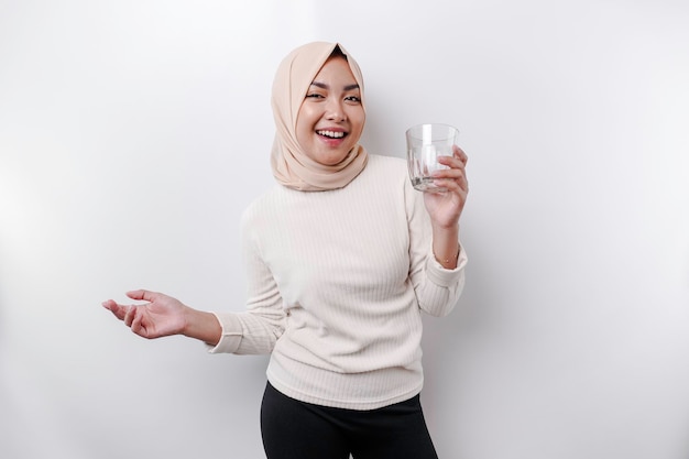 Una alegre mujer musulmana asiática con pañuelo en la cabeza está bebiendo un vaso de agua aislado de fondo blanco