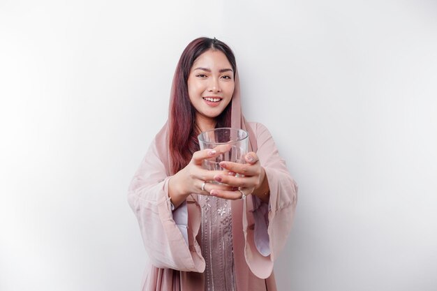 Una alegre mujer musulmana asiática con pañuelo en la cabeza está bebiendo un vaso de agua aislado de fondo blanco