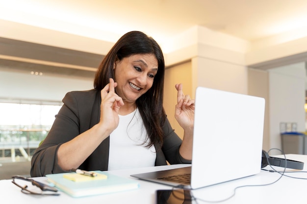 Alegre mujer de negocios asiática cruzando los dedos mientras se sienta en la computadora portátil en el interior de la oficina moderna