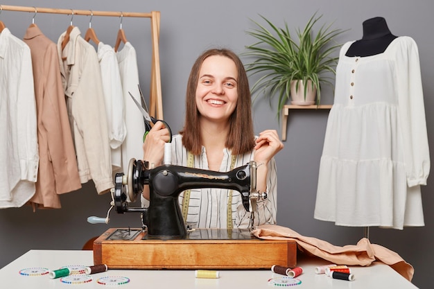 Alegre mujer de negocios caucásica modista trabajando en una máquina de coser en su propio taller de sastrería sosteniendo tijeras mirando la cámara con expresión facial positiva