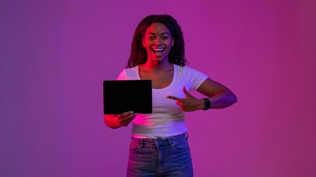 Alegre mujer negra apuntando a la tableta digital con pantalla en blanco en la mano