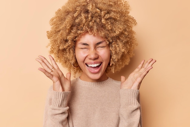 Alegre mujer optimista mantiene las palmas levantadas grita felizmente cierra los ojos smils ampliamente muestra dientes blancos vestido posada puente casual aislado sobre fondo de estudio marrón Concepto de reacciones humanas