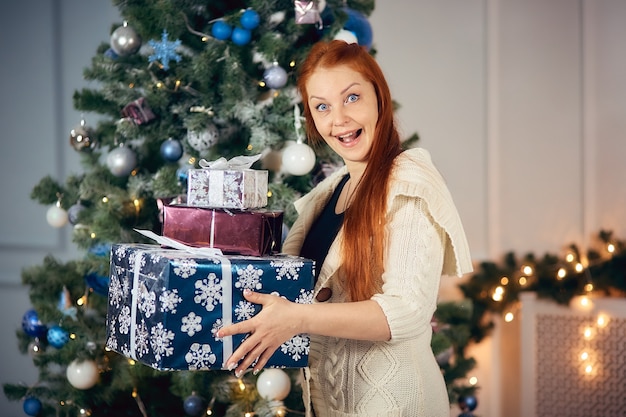 Alegre mujer pelirroja con un montón de regalos para Navidad cerca del árbol de Navidad