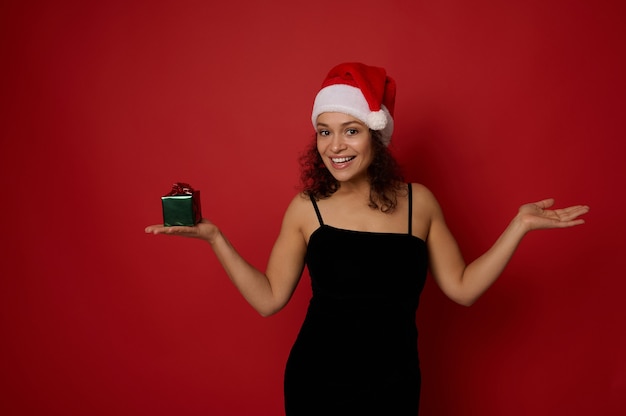 Alegre mujer de raza mixta que lleva un gorro de Papá Noel, sonríe con una sonrisa dentuda mirando a la cámara, sostiene un regalo de Navidad en una mano y levanta la otra mano con la palma hacia arriba sosteniendo un espacio de copia imaginaria sobre fondo rojo