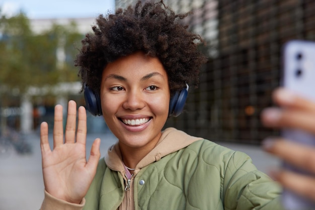 Alegre mujer rizada se toma un autorretrato a través de un teléfono inteligente con ondas, la palma dice hola, escucha música a través de auriculares, sonríe alegremente, posa al aire libre contra un fondo borroso, tiene recreación usando tecnología