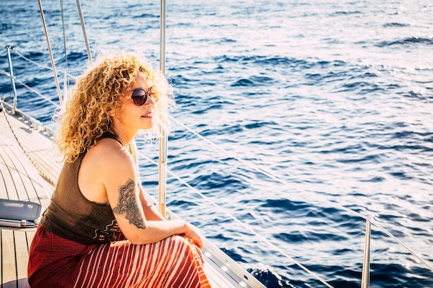 Alegre mujer rubia adulta joven atractiva sonrisa y disfrutar del viaje de excursión sentado en la cubierta del barco de vela mirando el mar azul alrededor