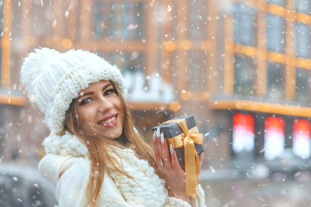 Alegre mujer rubia en bata blanca sosteniendo una caja de regalo en la calle durante las nevadas durante las nevadas. Espacio para texto