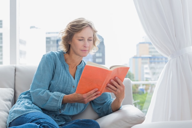 Alegre mujer rubia sentada en su sofá leyendo un libro