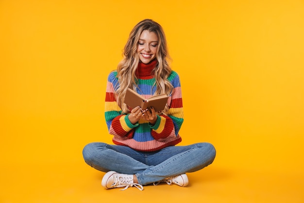 Alegre mujer rubia sonriendo y leyendo un libro mientras está sentado en el piso aislado sobre la pared amarilla