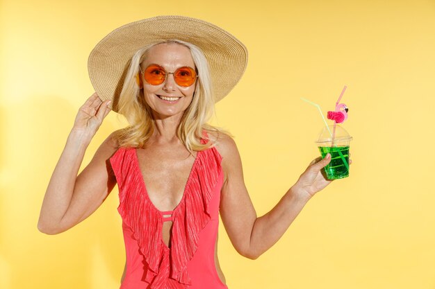 Alegre mujer rubia en traje de baño rojo gafas de sol y sombrero de paja sonriendo a la cámara ajustando su sombrero