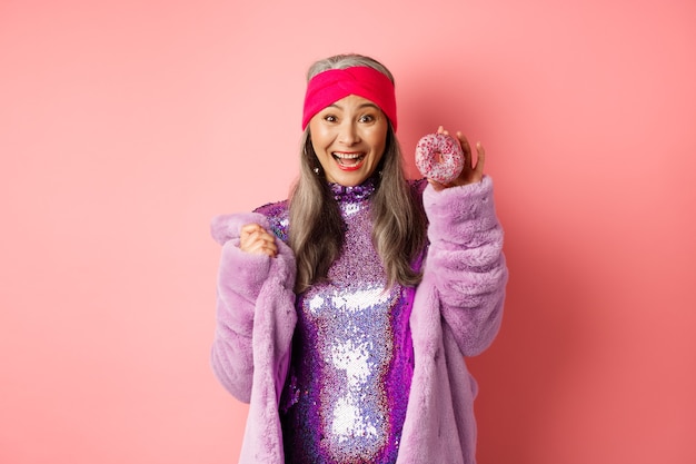 Alegre mujer senior asiática con cabello gris, vestido con purpurina de fiesta, mostrando una deliciosa rosquilla y sonriendo feliz, comiendo dulces, de pie sobre fondo rosa