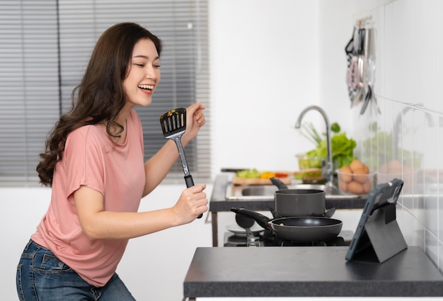 Alegre mujer vloguera cantando con turner y transmisión en vivo desde una tableta digital mientras cocina en la cocina en casa