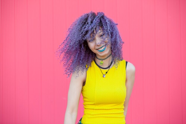 Alegre mulher adolescente com cabelo afro roxo e lábios azuis em uma parede rosa