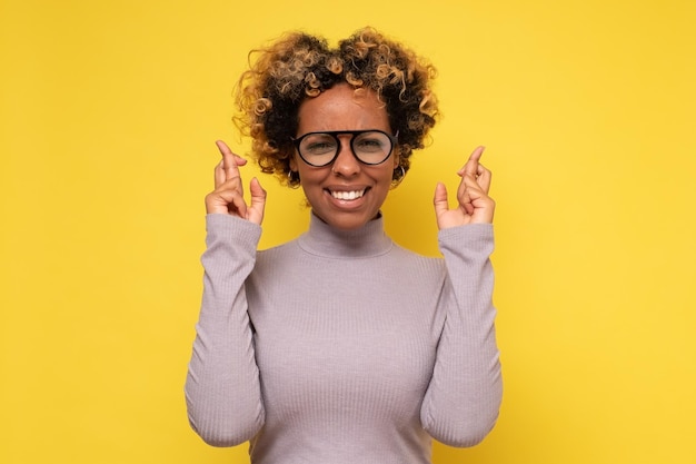 Alegre mulher afro-americana aguarda orações milagrosas e espera que os sonhos se tornem realidade cruza os dedos para dar boa sorte