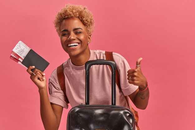 Alegre mulher afro-americana com bilhetes passaporte e mala em fundo rosa