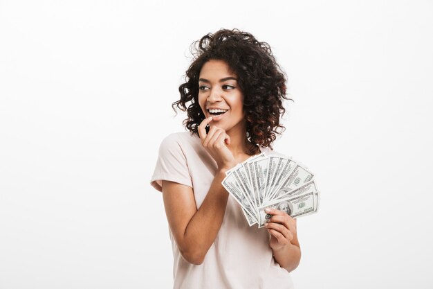 Alegre mulher americana com penteado afro e grande sorriso segurando leque de dinheiro dólar em dinheiro, isolado sobre a parede branca