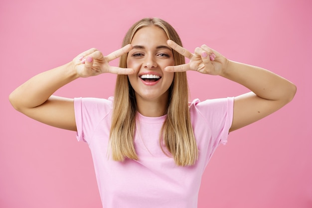 Alegre mulher amigável com cabelos loiros em t-shirt casual, mostrando paz contra um fundo rosa.